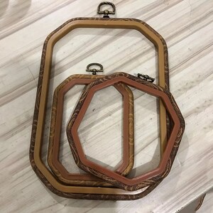 May include: Three octagonal embroidery hoops with wooden frames and brass clasps. The hoops are nested inside each other, with the largest hoop on the bottom and the smallest hoop on top.