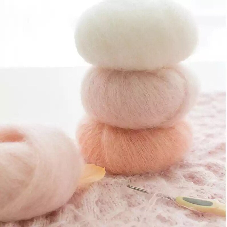May include: Four balls of yarn in different shades of pink, from white to a deep peach, stacked on top of each other. The yarn is fluffy and soft, and the balls are arranged in a pyramid shape.