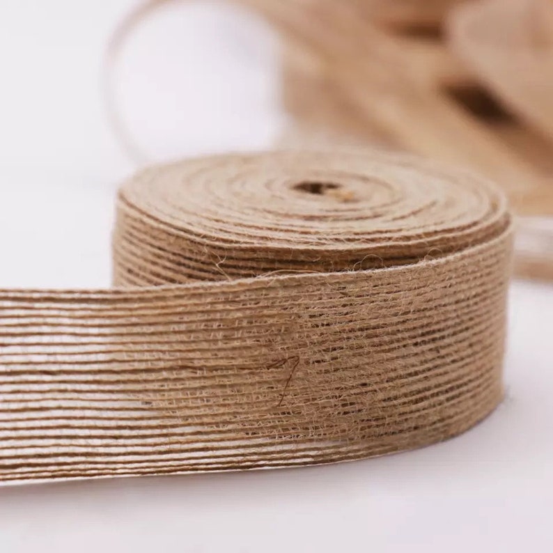 Op de afbeelding: Een rol natuurlijke bruine juteband met een getextureerd, geweven ontwerp. Het lint is opgerold en het uiteinde is uitgerold, waardoor de textuur van het materiaal zichtbaar wordt.