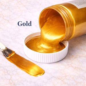 Gold metallic acrylic paint pouring from a jar into a lid with a matching gold brushstroke, shown in a square product image on a soft marble background.