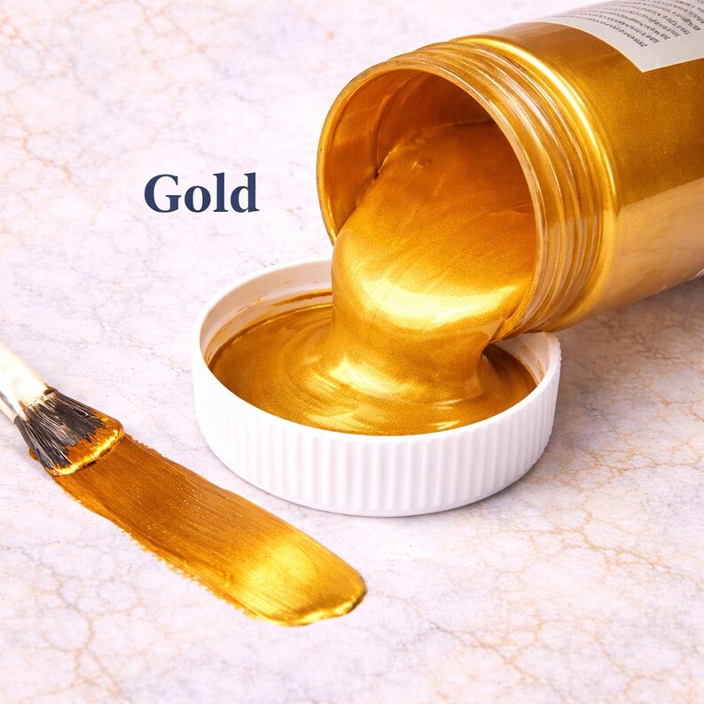 Gold metallic acrylic paint pouring from a jar into a lid with a matching gold brushstroke, shown in a square product image on a soft marble background.