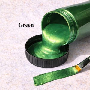 Copper green metallic acrylic paint pouring from a jar into a lid with a matching copper green brushstroke, shown in a square product image on a soft marble background.