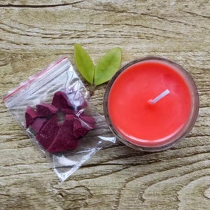 Puede incluir: Una vela en un tarro de vidrio transparente con cera de color naranja brillante y una mecha blanca. Una peque&ntilde;a bolsa de pl&aacute;stico con trozos de cera roja est&aacute; al lado de la vela.