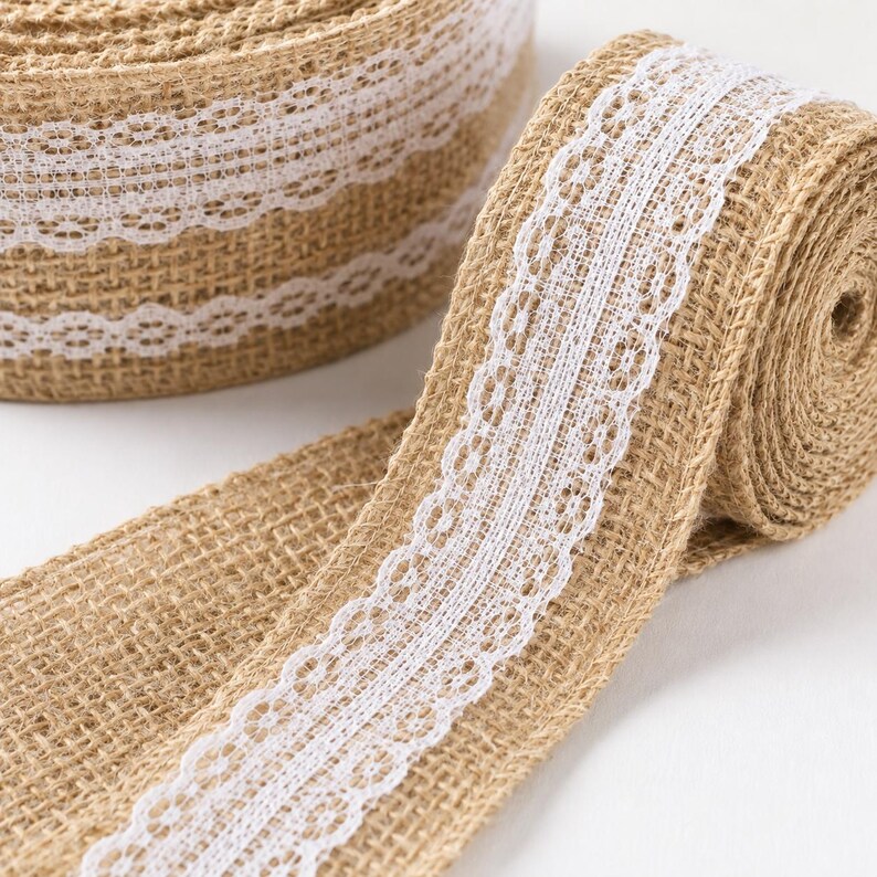 May include: A roll of tan burlap ribbon featuring a centered white lace trim with a delicate floral pattern. The coarse, woven texture of the natural brown fabric contrasts with the smooth, intricate white lace detailing along the center.