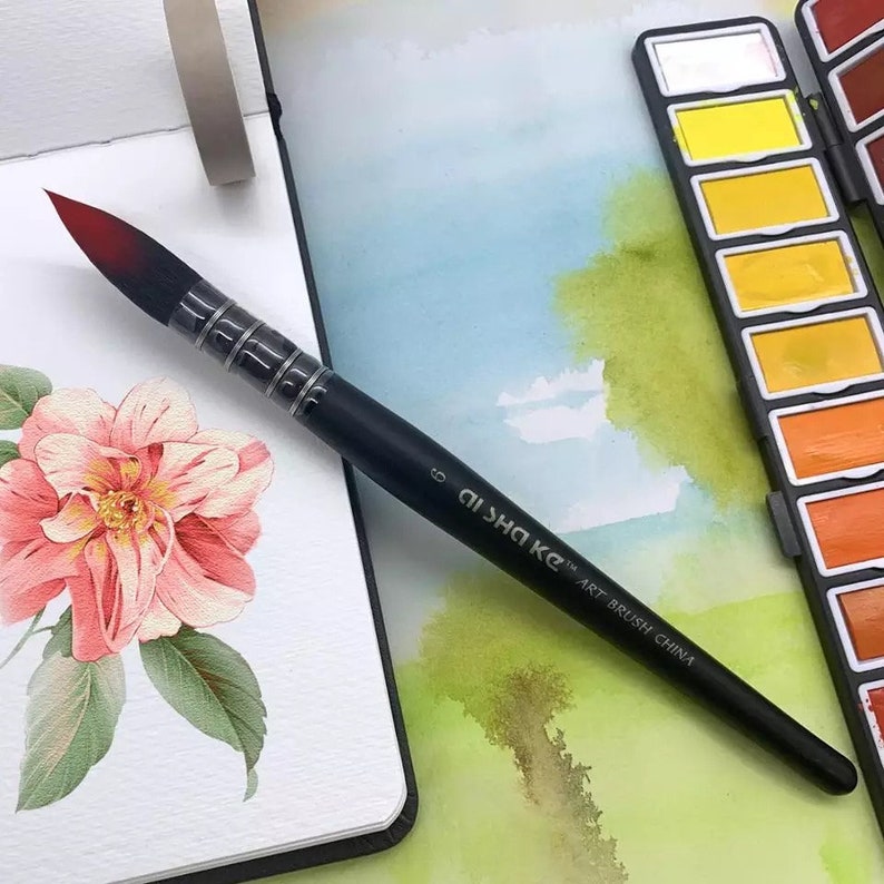 Op de afbeelding: Een zwarte ronde aquarelkwast met een donkerrode punt, voorzien van de tekst AISHA KC ART BRUSH CHINA en het cijfer 6 op de steel. De kwast heeft een zilveren huls en ligt naast een aquarelverfset en een schetsboek.