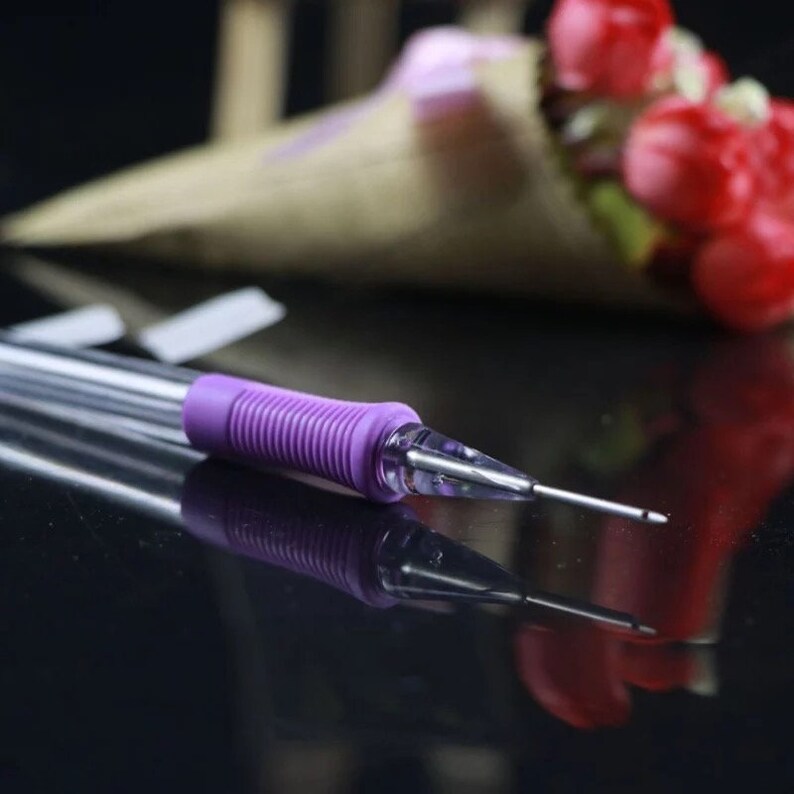 May include: A close-up of a purple and clear plastic embroidery needle with a sharp point. The needle is lying on a black reflective surface.