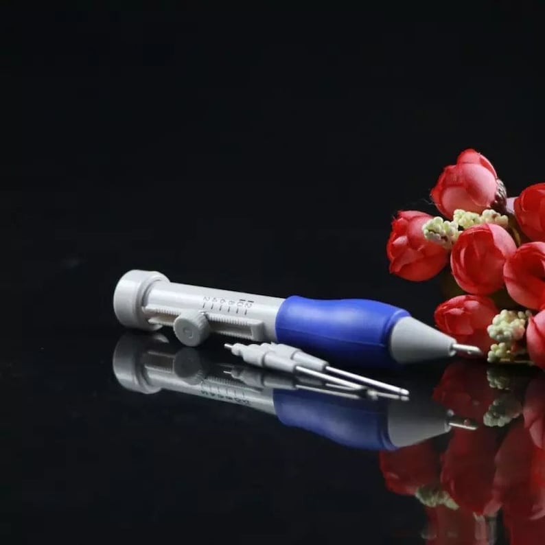 May include: A blue and white plastic needle threader tool with a needle guide and a knob to adjust the needle depth. The tool is on a black surface with a bouquet of red roses in the background.