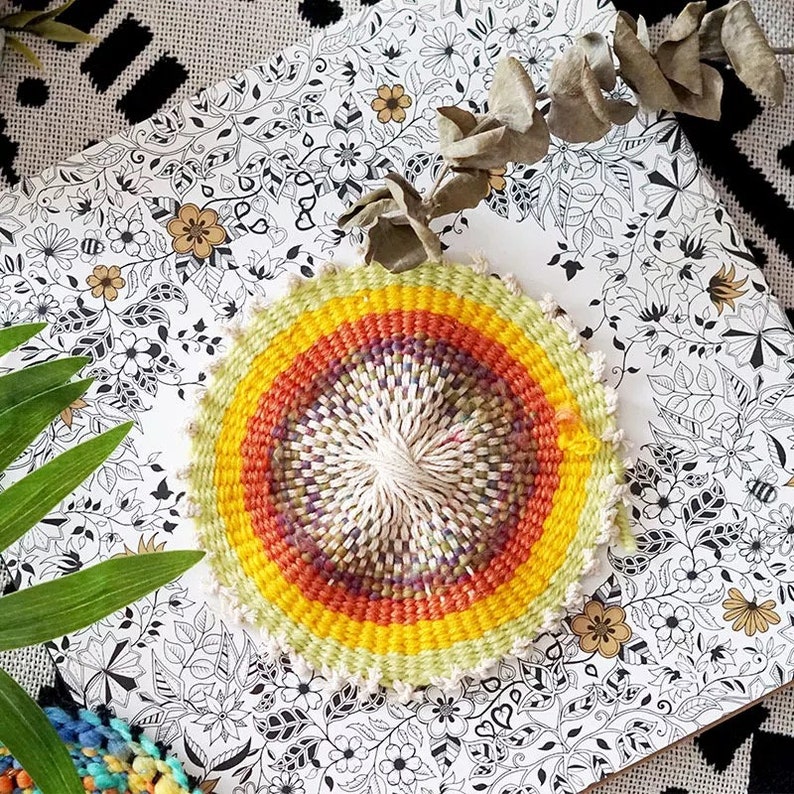 May include: A circular woven wall hanging with a rainbow pattern of yellow, orange, red, purple, and green. The center of the hanging is white and the outer edge is a light green.