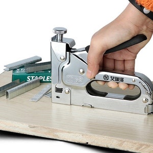 May include: A silver staple gun with a black handle is held by a person's hand. Several silver staples and a green box of staples are next to the staple gun. The staple gun is on a light brown wooden surface.