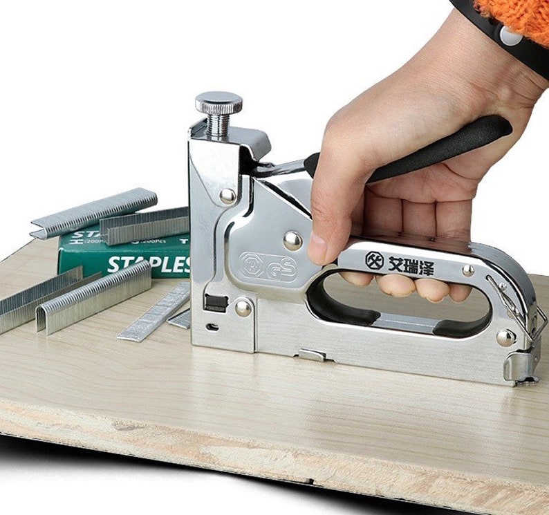 May include: A silver staple gun with a black handle is held by a person's hand. Several silver staples and a green box of staples are next to the staple gun. The staple gun is on a light brown wooden surface.