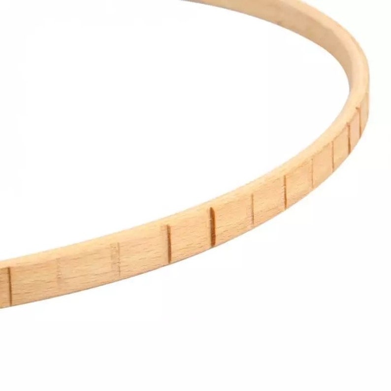 May include: A close-up of a wooden embroidery hoop with a natural finish. The hoop is made of smooth, light-colored wood and has a series of evenly spaced grooves around the edge.