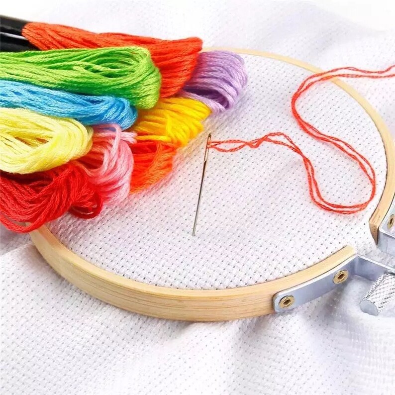 Puede incluir: Primer plano de un aro de bordado de madera con tela Aida blanca estirada sobre &eacute;l. Una aguja y hilo de bordar rojo est&aacute;n enhebrados en la tela. Un paquete de hilo de bordar de colores est&aacute; colocado al lado del aro.