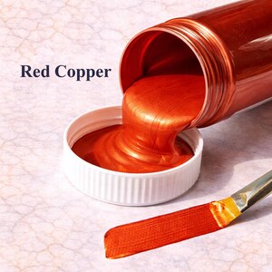 Red copper metallic acrylic paint pouring from a jar into a lid with a matching red copper brushstroke, shown in a square product image on a soft marble background.