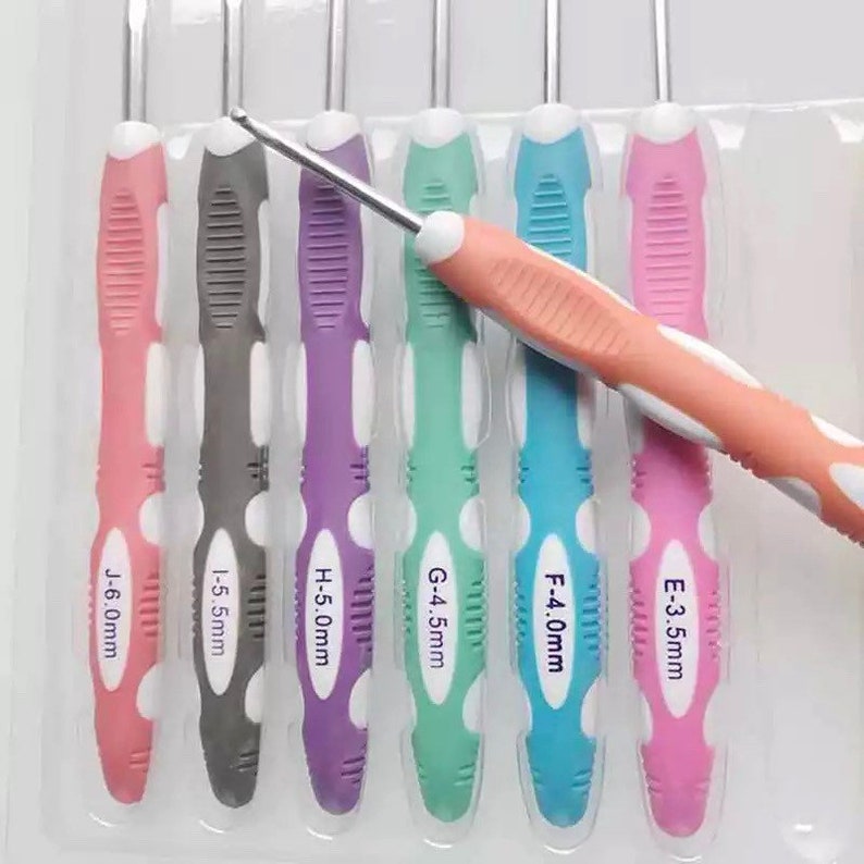 May include: A set of six crochet hooks with ergonomic handles in pink, gray, purple, mint green, blue, and light pink. Each handle features white accents and is printed with size labels ranging from E-3.5mm to J-6.0mm, arranged in a clear tray.