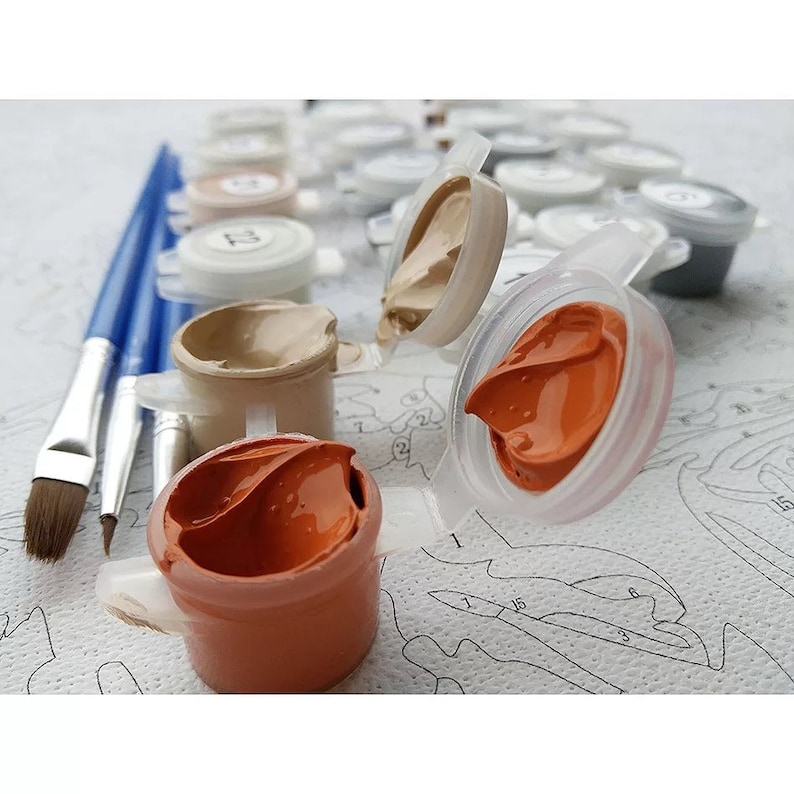 May include: A paint-by-number kit featuring a set of small, connected plastic pots filled with various shades of acrylic paint, including orange and beige. Three blue-handled paintbrushes lie beside the open pots on a printed canvas.