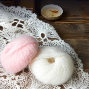 May include: Two balls of yarn, one pink and one white, are displayed on a white crocheted surface. The yarn is soft and fluffy, perfect for knitting or crocheting.