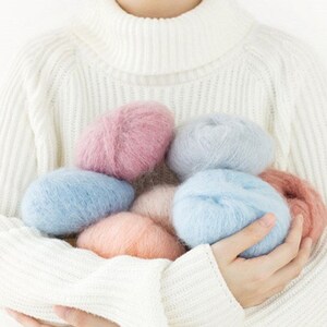 May include: A collection of soft, fluffy yarn in shades of pink, blue, and peach. The yarn is arranged in a pile and held in the hands of a person.