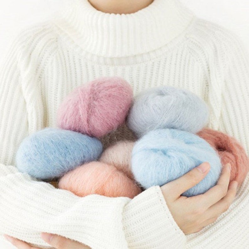 May include: A collection of soft, fluffy yarn in shades of pink, blue, and peach. The yarn is arranged in a pile and held in the hands of a person.