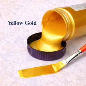 Yellow gold metallic acrylic paint pouring from a jar into a lid with a matching yellow gold brushstroke, shown in a square product image on a soft marble background.