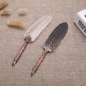 Pode incluir: Duas canetas de pena de estilo antigo com pontas de metal ornamentadas e penas de pena. Uma pena &eacute; branca com listras pretas, a outra &eacute; marrom com manchas pretas.