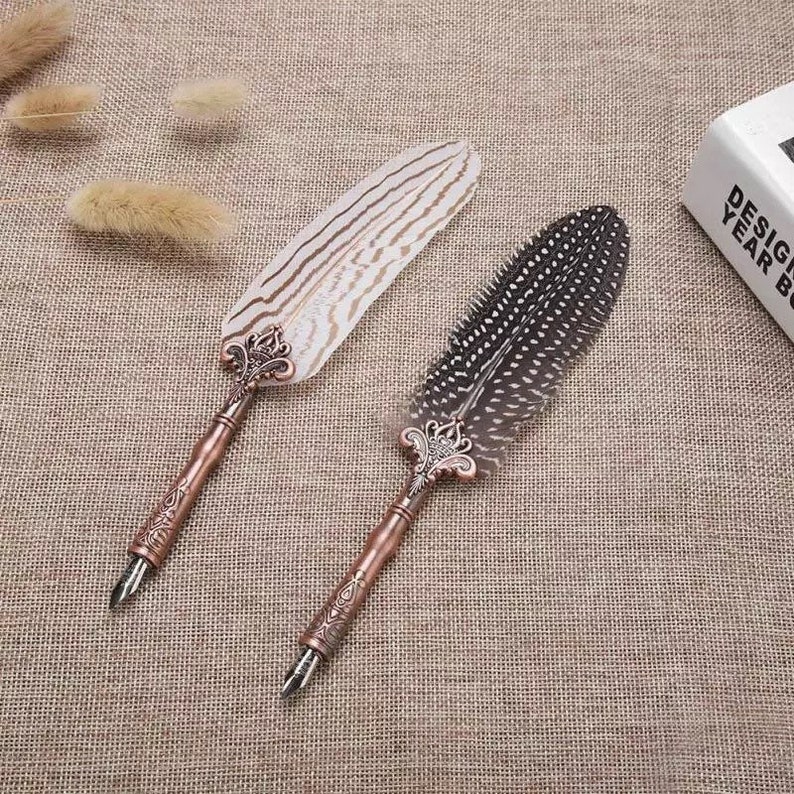 Pode incluir: Duas canetas de pena de estilo antigo com pontas de metal ornamentadas e penas de pena. Uma pena &eacute; branca com listras pretas, a outra &eacute; marrom com manchas pretas.