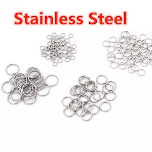 Puede incluir: Una colección de pequeños y brillantes anillos de acero inoxidable. Los anillos son de color plateado y están dispersos sobre un fondo blanco. El texto "Stainless Steel" se muestra en rojo en la parte superior de la imagen.