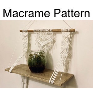 May include: White macrame wall hanging with a wooden dowel and a shelf. The macrame is in a diamond pattern and has a plant in a clear glass vase on the shelf.