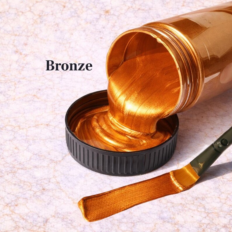 Bronze metallic acrylic paint pouring from a jar into a lid with a matching bronze brushstroke, shown in a square product image on a soft marble background.