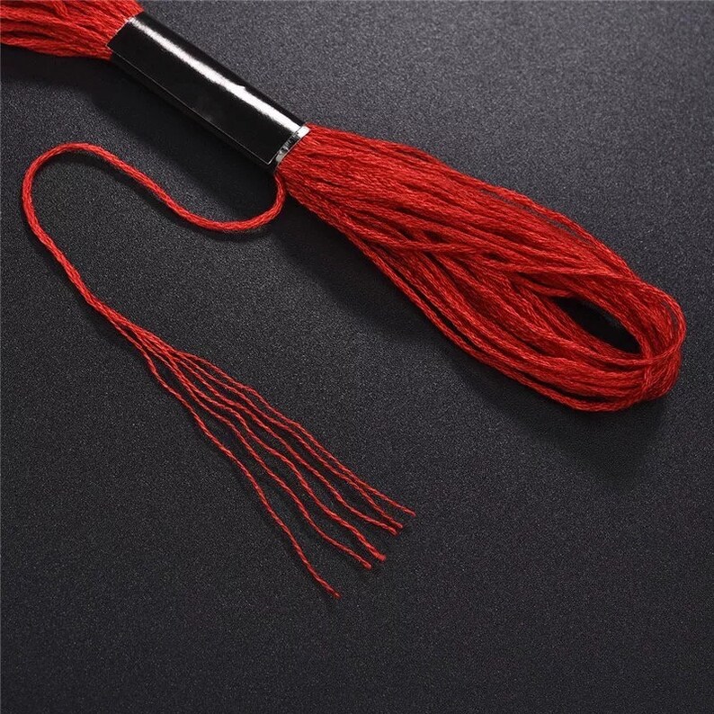 Puede incluir: Una bobina de hilo de bordar rojo con una bobina de pl&aacute;stico negro. El hilo est&aacute; desenrollado y colgando, creando un efecto de flecos.