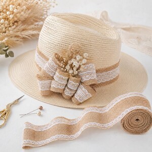 May include: A beige straw fedora hat featuring a wide band of burlap ribbon with white lace trim. The hat is decorated with a cluster of dried white flowers and a burlap bow. A roll of matching burlap ribbon and gold scissors are nearby.
