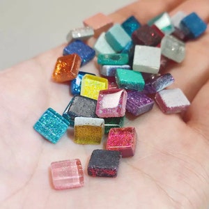 Puede incluir: Una colección de pequeñas baldosas cuadradas y brillantes de varios colores, incluyendo azul, verde, rojo, amarillo y morado. Las baldosas están dispuestas en una pila sobre una mano.