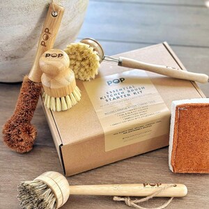 May include: A collection of kitchen cleaning tools, including brushes, a sponge, and a box set. The brushes have wooden handles and natural bristles. The box set is labeled "Essential Kitchen Brush Starter Kit" with the brand name "POP".