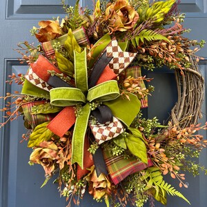 May include: A fall-themed wreath with a woven grapevine base. It features faux flowers in shades of brown and orange, green leaves, and various ribbons in plaid, solid orange, and black. The wreath is displayed on a blue door.