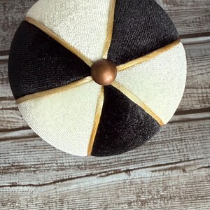 May include: A round, decorative object with a segmented design. Alternating sections of black and off-white fabric are separated by gold-colored trim. A small, copper-colored sphere is at the center.