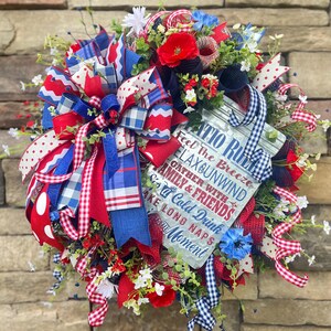 May include: A patriotic wreath with red, white, and blue ribbons, flowers, and greenery. A sign with the words "PATIO RULES" and other text is attached to the wreath. The wreath is displayed against a stone wall.