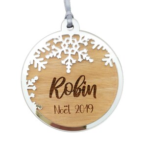 Peut inclure: Une boule de Noël ronde en bois avec un bord argenté. La boule présente un motif de flocon de neige blanc et le nom "Robin" gravé sur le bois. Le texte "Noël 2019" est également gravé sur la boule.
