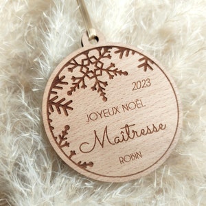 Peut inclure: Une décoration de Noël ronde en bois avec un motif de flocon de neige, le texte "Joyeux Noël 2023" et le mot "Maîtresse" écrit en cursive. La décoration comprend également le nom "Robin" en bas.