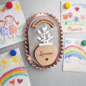 Peut inclure: Un aimant encadré en or rose avec un motif de plante en bois. L'aimant porte les inscriptions "MAMIE D'AMOUR" et "Louise". L'aimant est affiché sur un réfrigérateur avec des dessins d'enfants.