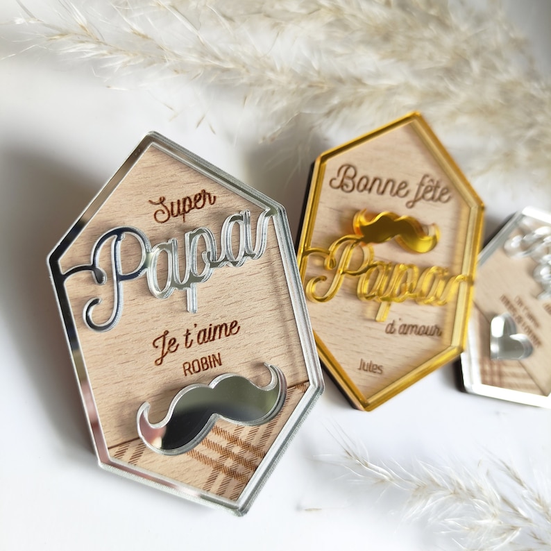 May include: Three wooden plaques with mirrored surfaces. The plaques are shaped like hexagons and feature the words "Super Papa" with a silver mirrored mustache, "Bonne fete Papa" with a gold mirrored mustache, and "Papa" with a silver mirrored heart. The plaques are perfect for Father's Day or a birthday.