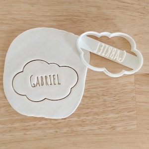 Può includere: Tagliapasta in plastica bianca a forma di nuvola con il nome "GABRIEL" scritto all'interno. Il tagliapasta è sopra un pezzo di pasta bianca con lo stesso disegno tagliato.