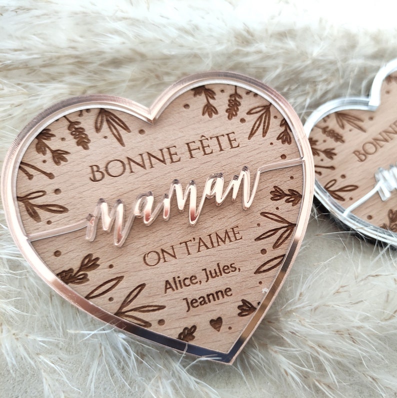 Puede incluir: Una placa de madera en forma de coraz&oacute;n con un borde de oro rosa. La placa tiene un dise&ntilde;o floral y el texto "BONNE F&Ecirc;TE maman ON T'AIME Alice, Jules, Jeanne" est&aacute; grabado en ella.