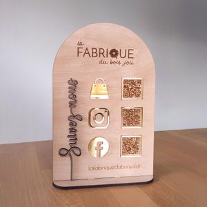 Könnte beinhalten: Ein Holzständer mit dem Text "la FABRIQUE du bois joli" und Social-Media-Symbolen. Der Ständer zeigt eine Einkaufstasche, Instagram- und Facebook-Symbole sowie QR-Codes zum Scannen. Die Worte "Suivez nous" sind ebenfalls angezeigt.
