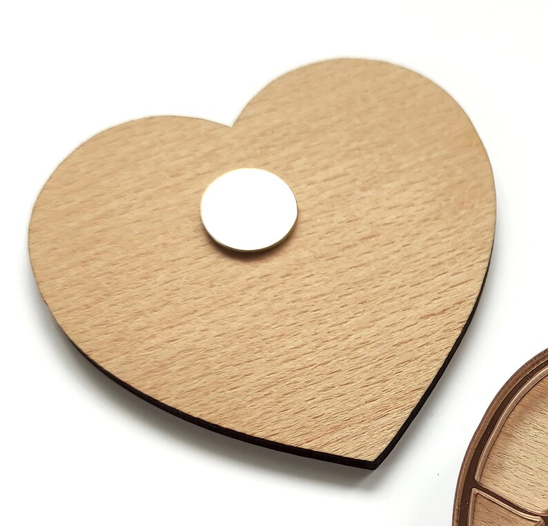 Puede incluir: Un adorno de madera en forma de coraz&oacute;n con un im&aacute;n redondo blanco pegado en la parte delantera.