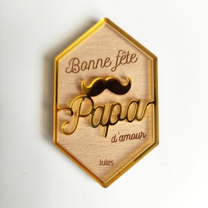 May include: A hexagonal wooden sign with a gold border and gold lettering. The sign says "Bonne f&ecirc;te Papa d'amour Jules". There is a gold glitter mustache above the word "Papa".