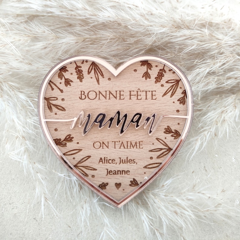 Puede incluir: Una placa de madera en forma de coraz&oacute;n con un borde de oro rosa. La placa tiene un dise&ntilde;o floral y el texto "Bonne F&ecirc;te Maman On T'aime Alice, Jules, Jeanne".