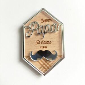 May include: A silver hexagonal frame with a wood background. The frame has the words "Super Papa" and "Je t'aime Robin" engraved on it. A black and silver mirrored mustache is at the bottom of the frame.