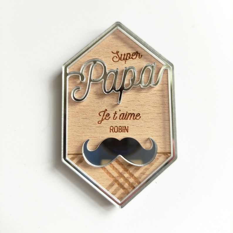 May include: A silver hexagonal frame with a wood background. The frame has the words "Super Papa" and "Je t'aime Robin" engraved on it. A black and silver mirrored mustache is at the bottom of the frame.