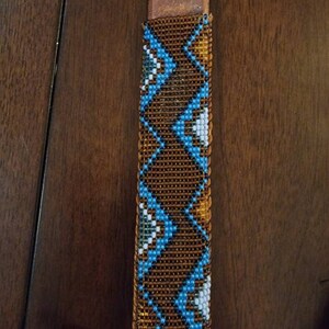 May include: Close-up of a brown leather strap with a beaded design. The beadwork features a geometric pattern in shades of blue, white, and gold against a brown background. The strap is likely part of a larger item, such as a belt or bag.