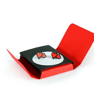 Może przedstawiać: Czerwone kolczyki w kształcie serca prezentowane w otwartym, czerwono-czarnym pudełku prezentowym. Kolczyki znajdują się na białym owalu, na czarnym tle. Pudełko jest otwarte, ukazując kolczyki.