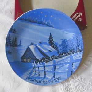 Può includere: Un piatto da collezione rotondo in porcellana raffigurante un paesaggio invernale blu e bianco con una baita innevata, una staccionata in legno e alberi di pino sotto un cielo stellato. Il testo WEIHNACHTEN 1977 è stampato in nero sul bordo.